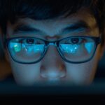 Close-up of a marketer’s thoughtful face illuminated by a computer screen showing user comments with question marks and glowing lightbulbs reflected in glasses — symbolizing insight and discovery from audience feedback.